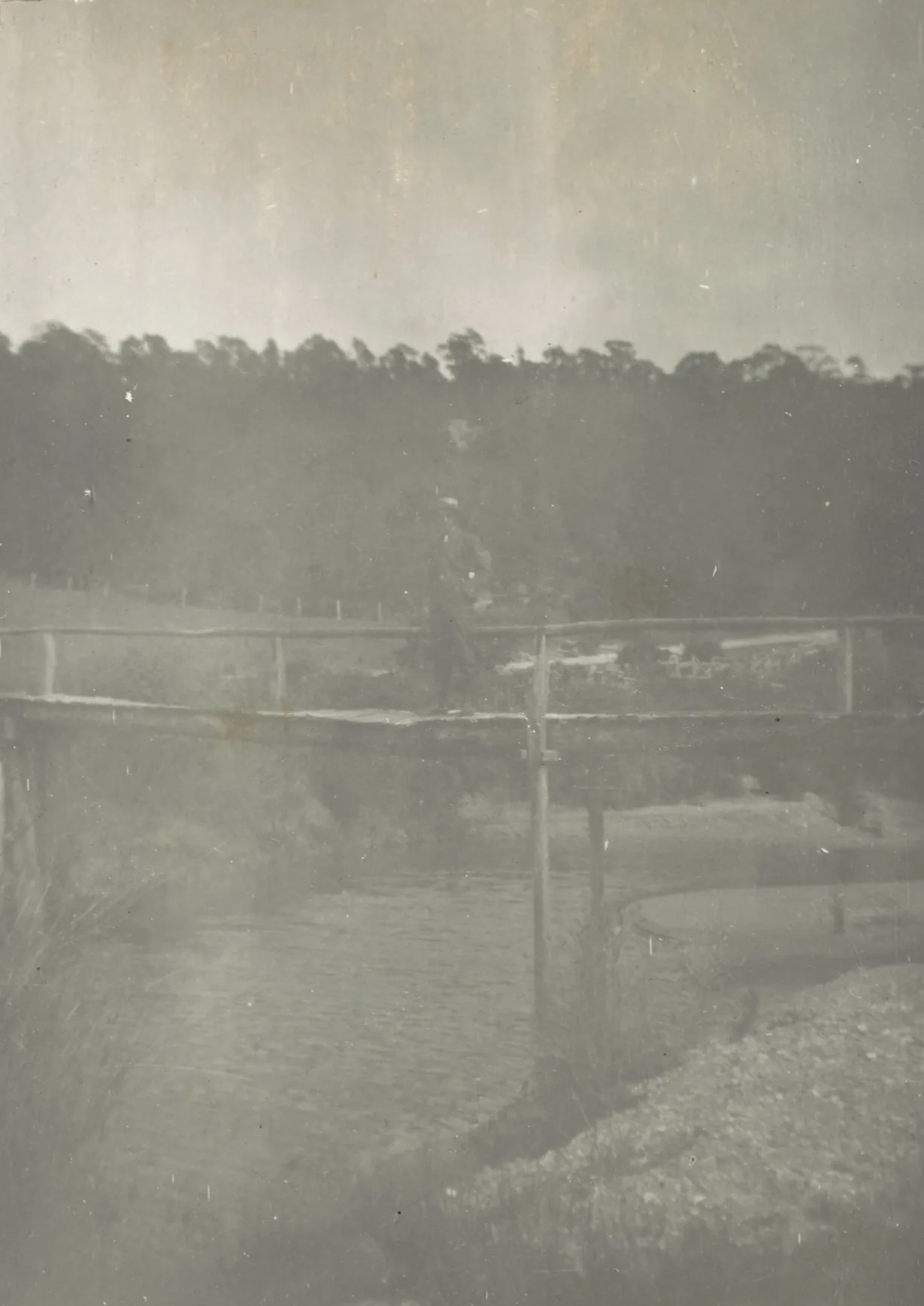 A faded snapshot of a man standing on a rough timber footbridge with a pole handrail over a still creek pool, reeds on one bank and gravel on the other, a fenced paddock and a dark line of bush behind.