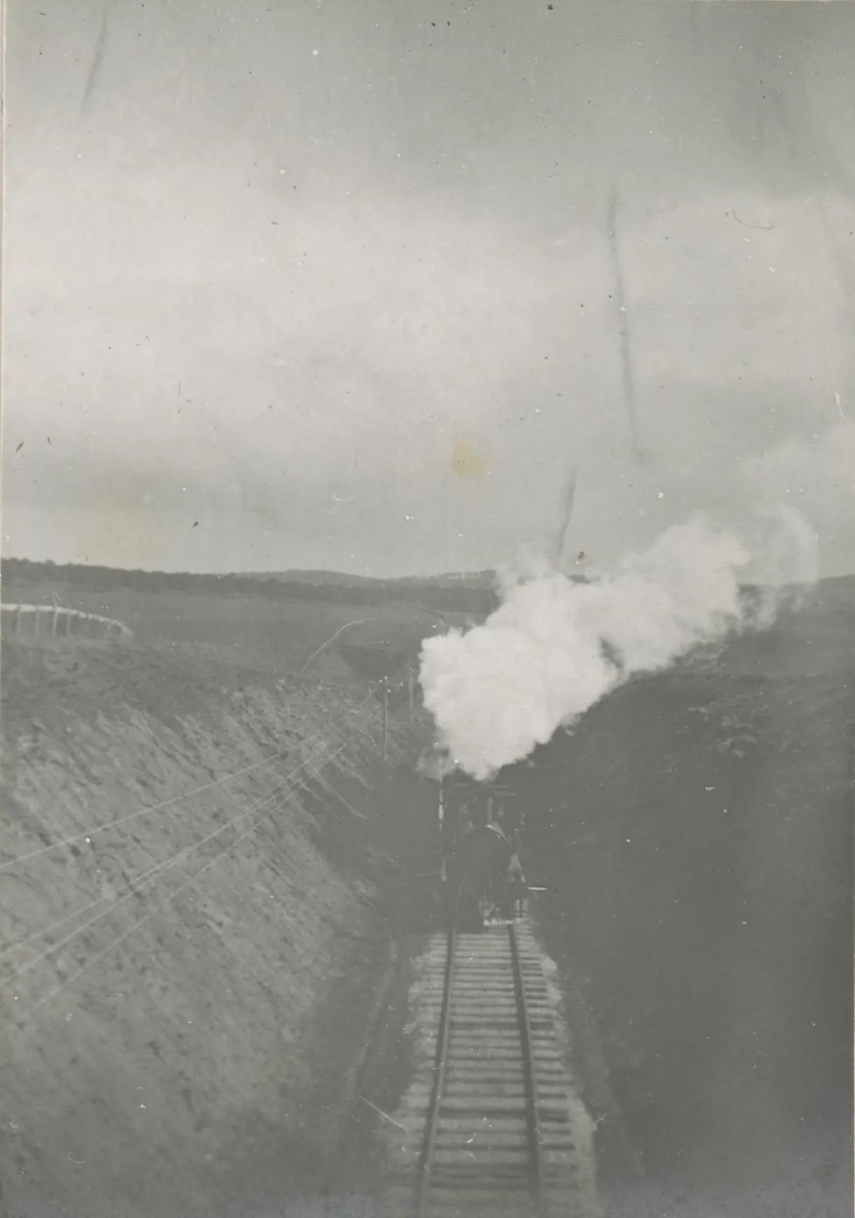 A steam engine coming head-on up a single track through a deep cutting, a white plume of steam rising over the paddocks.