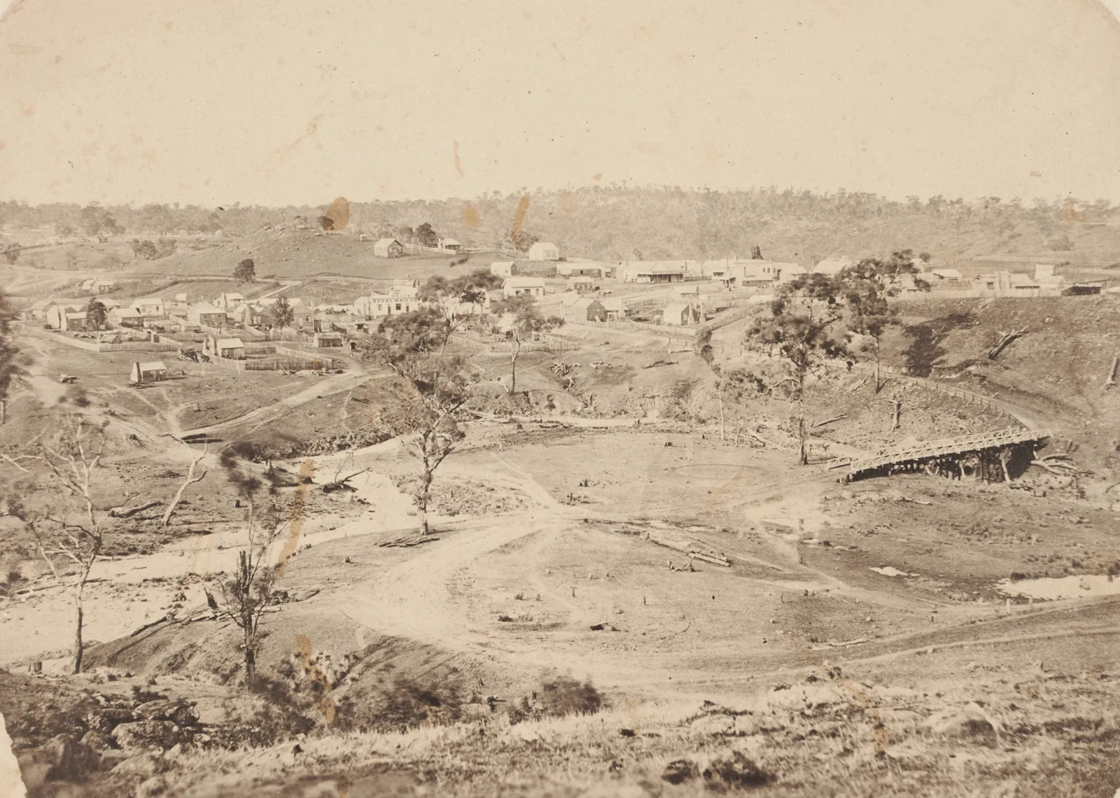 A bare, dug-over river flat winding across the middle of the picture, a timber crossing over the gully on the right, and the weatherboard houses and stores of Piggoreet spread over the rise behind, with ringbarked trees and bush-covered hills beyond.